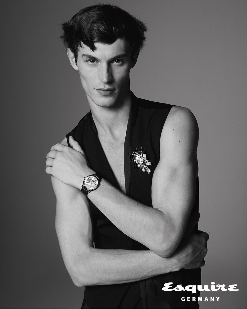 Black and white portrait of a slim male fashion model posing in a sleeveless top with a decorative pin and wristwatch against a grey background.