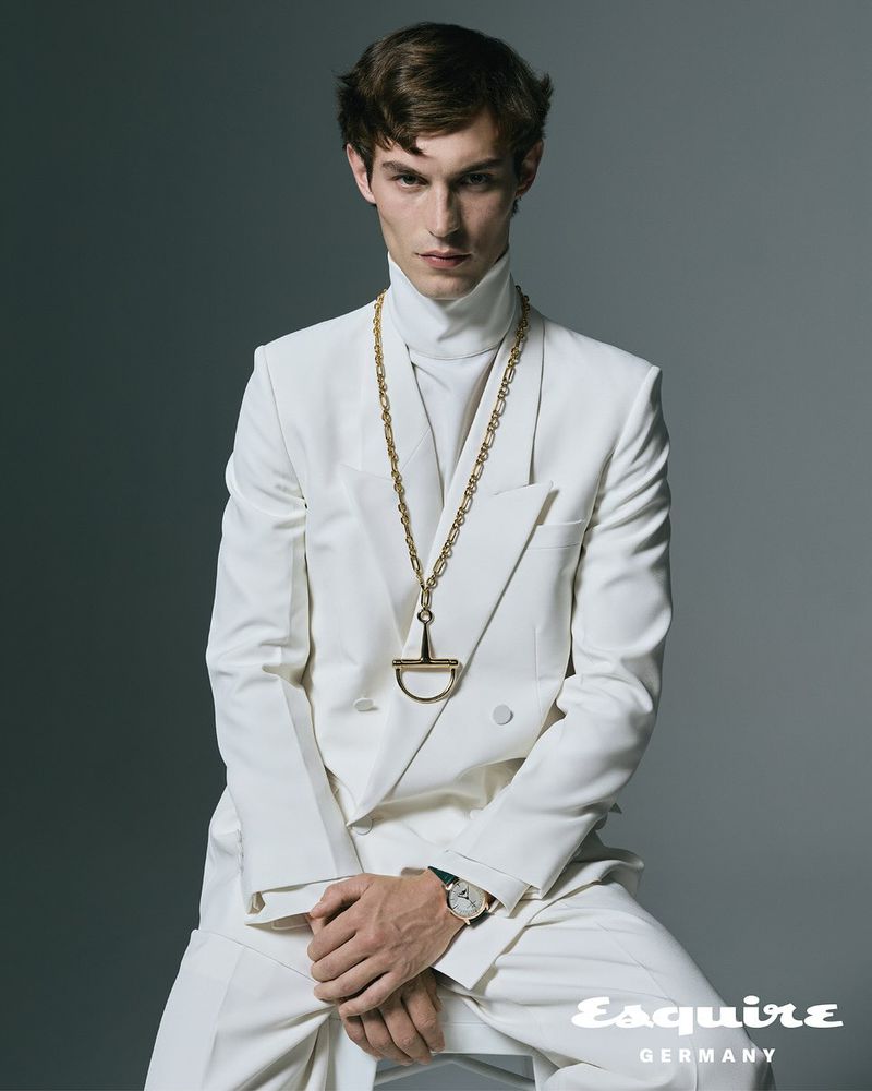 Portrait of a young adult male wearing a white suit with a gold chain necklace and watch posing in a photo studio.