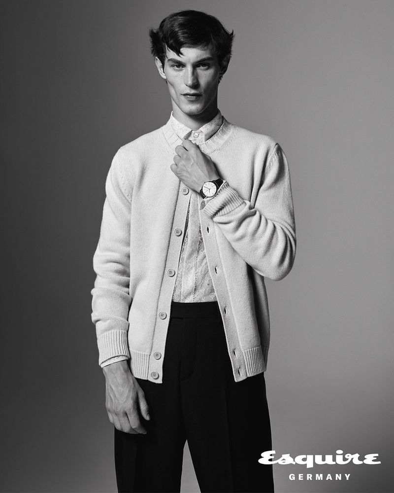 Black-and-white studio shot of a young adult male model wearing a button-down shirt, cardigan, and trousers while posing and looking at the camera.