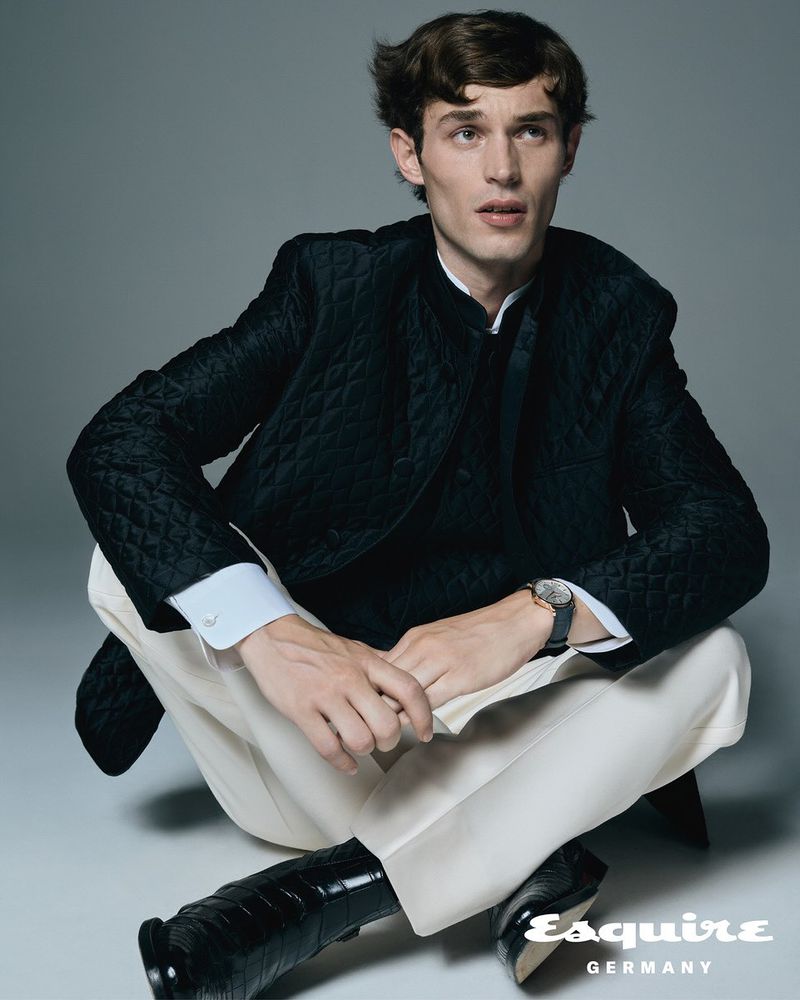 A single adult male is sitting cross-legged on a grey backdrop wearing a quilted black jacket, white pants, and black patent leather shoes.