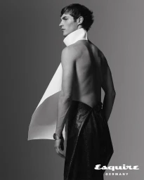 Kit Butler in black-and-white fashion portrait wearing a high-collared white wrap and dark trousers against a plain grey background