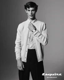 Model Kit Butler wearing a knit cardigan and dark tailored trousers, touching his collar against a studio background.