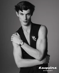 Monochrome portrait of Kit Butler wearing a sleeveless black top with a brooch and a wrist watch against a grey background.