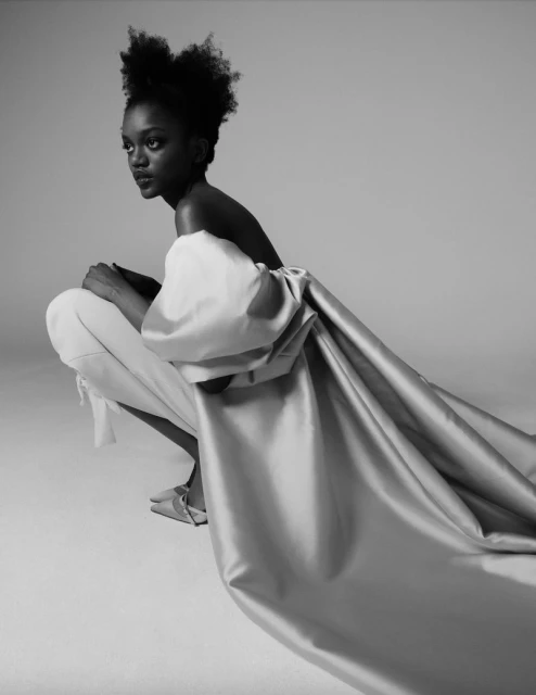 Black and white fashion portrait of an African woman with natural hair in flowing satin top and pants squatting in a photo studio.