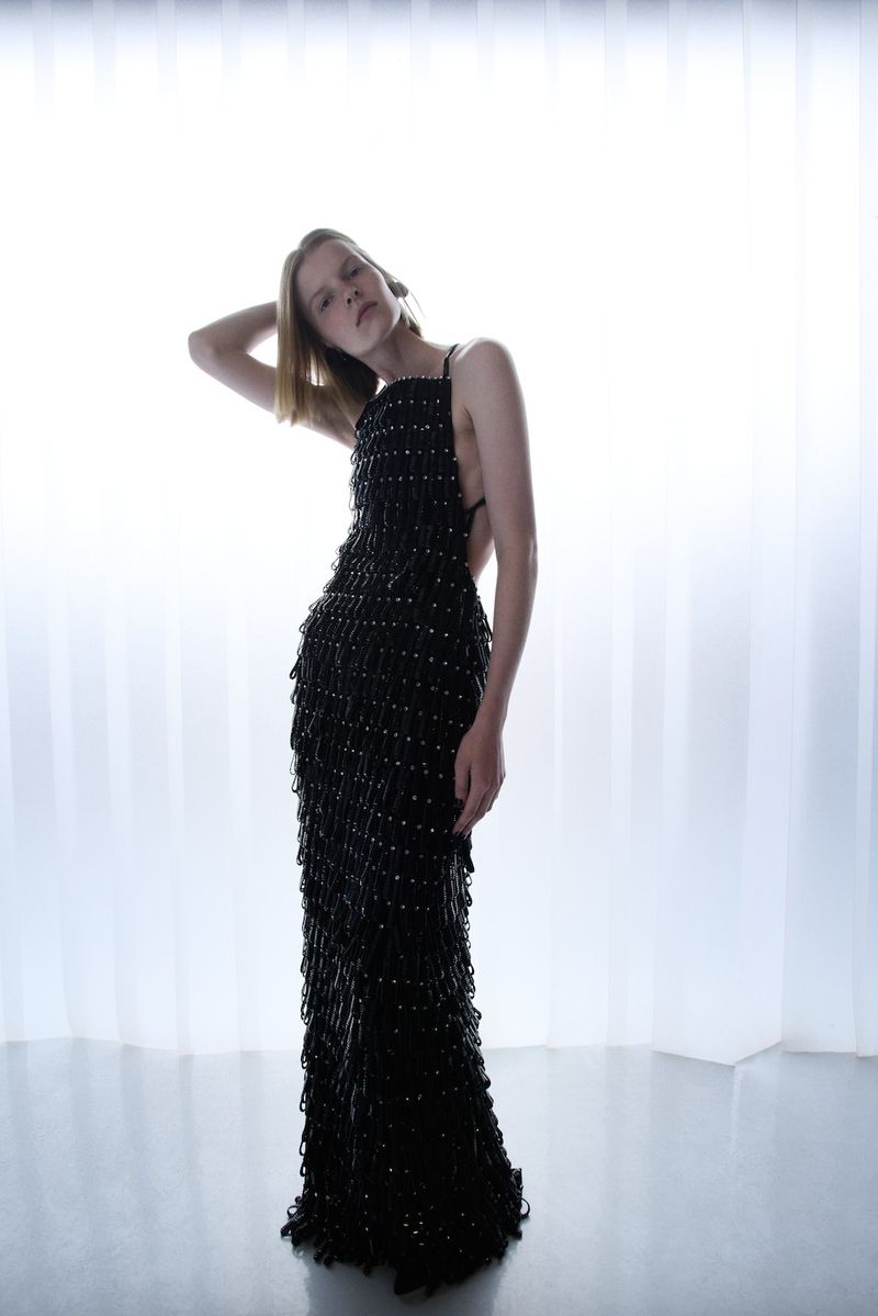 Adult female posing in full-length black beaded gown against a bright white background in a fashion studio with reflective floor.