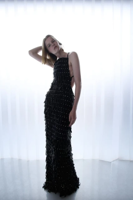 Adult female posing in full-length black beaded gown against a bright white background in a fashion studio with reflective floor.