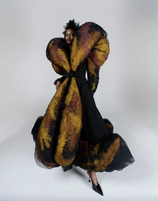 Fashion model in an oversized black couture dress with amber and brown accents poses in a photo studio against a white backdrop.