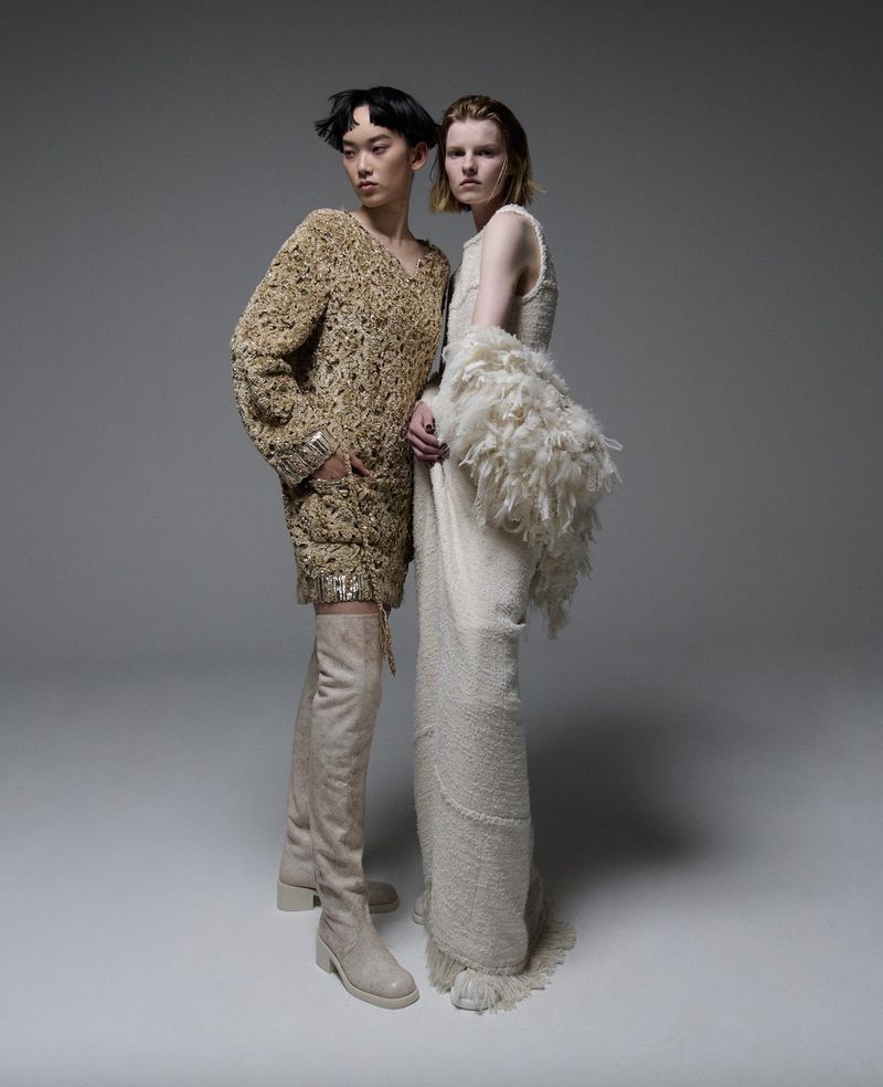 Two fashion models in studio setting. One wears beige thigh-high boots with a heavily textured minidress; the other in a long dress.