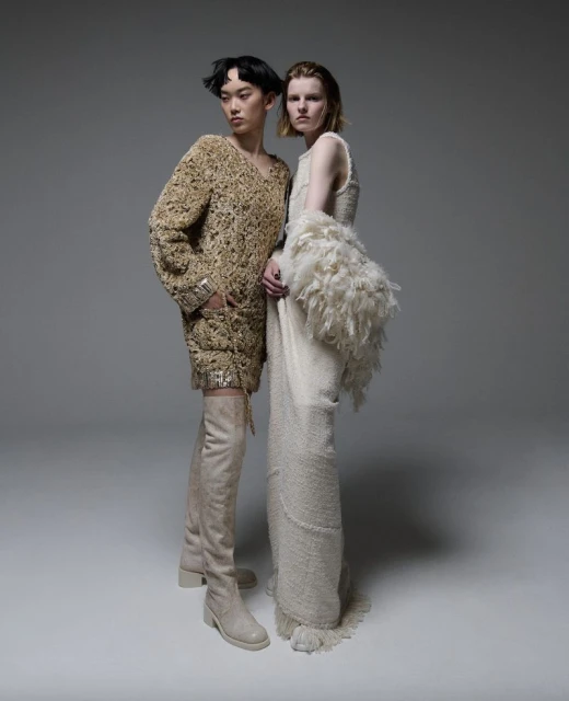 Two fashion models in studio setting. One wears beige thigh-high boots with a heavily textured minidress; the other in a long dress.