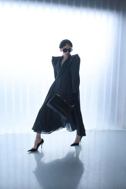 A woman in a black and white polka dot dress and black heels strikes a pose against a bright white backdrop, holding a black purse.