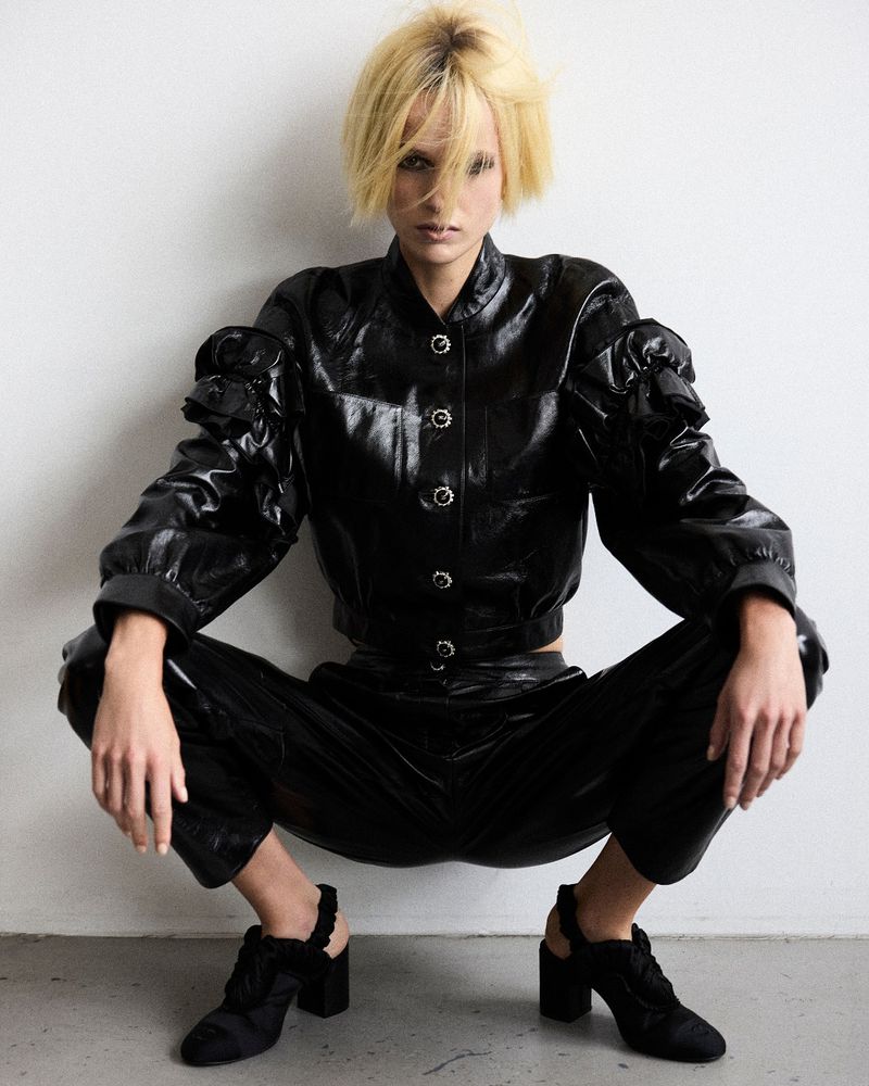 Stylish woman crouches against white wall, wearing black patent leather-like jacket and pants, with blonde bob haircut, high heel shoes.