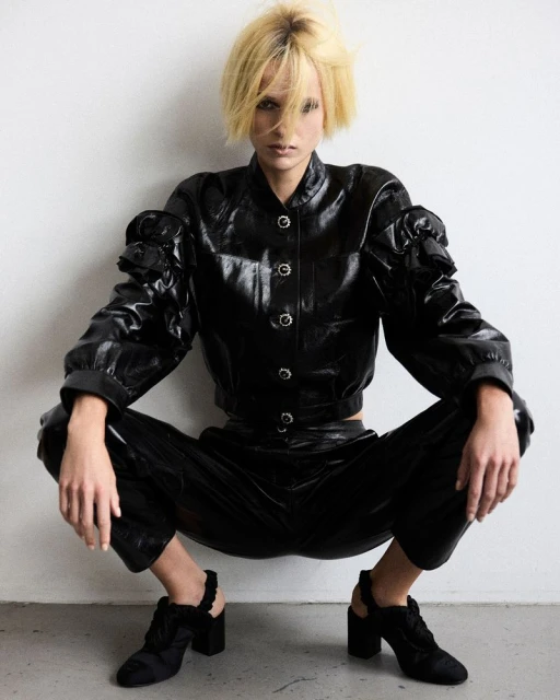 Stylish woman crouches against white wall, wearing black patent leather-like jacket and pants, with blonde bob haircut, high heel shoes.