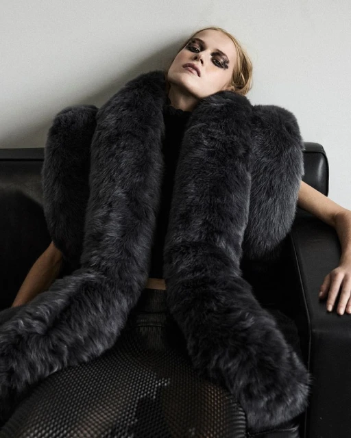 A woman with dramatic eye makeup poses on a black couch wearing a large grey faux fur stole, black top, and skirt with patterned mesh.