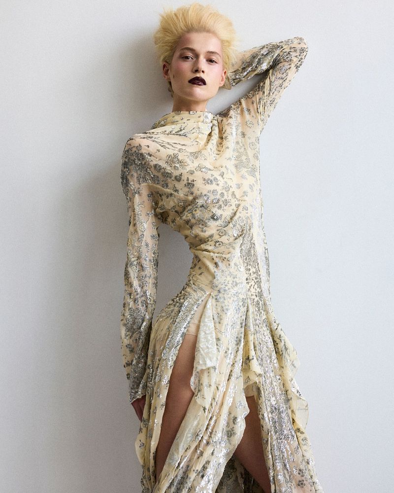 Woman in flowing gown with long sleeves and thigh-high slit, leaning against white wall, dark lipstick and blonde hair, fashion photography.