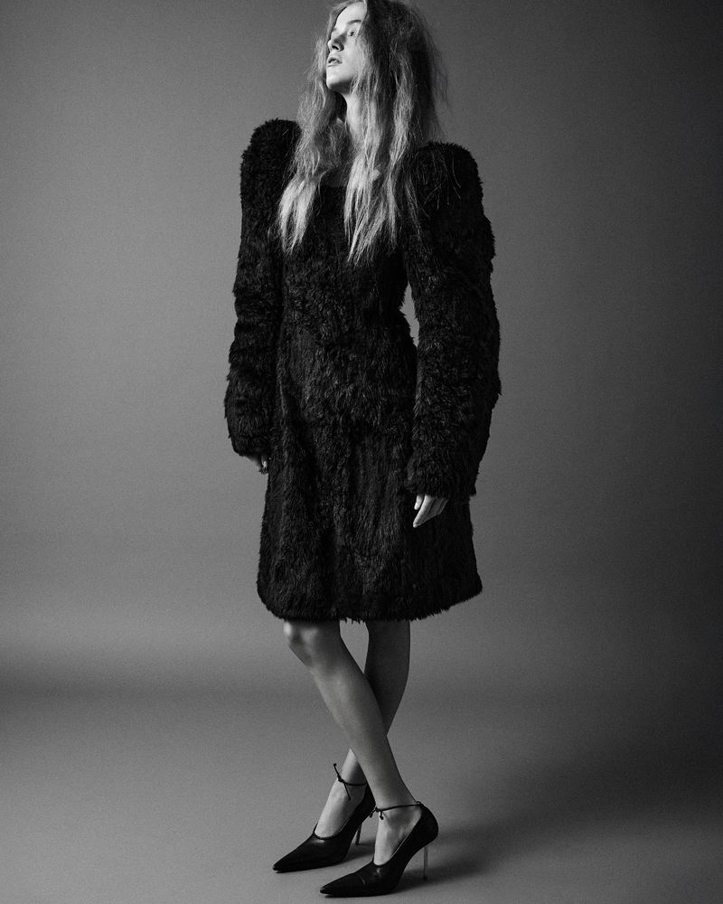 Monochrome studio shot of a woman in a black fur coat and heels, standing against a gray backdrop, looking off-camera.