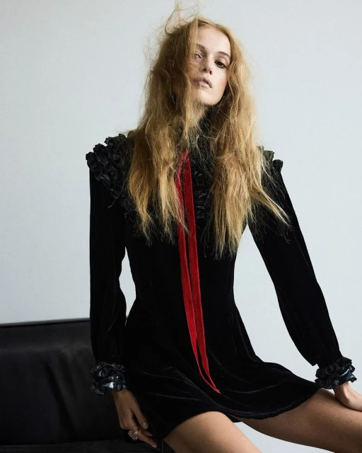 Portrait of a blonde woman in a black velvet dress with a red ribbon, seated indoors against a neutral background, fashion photography.