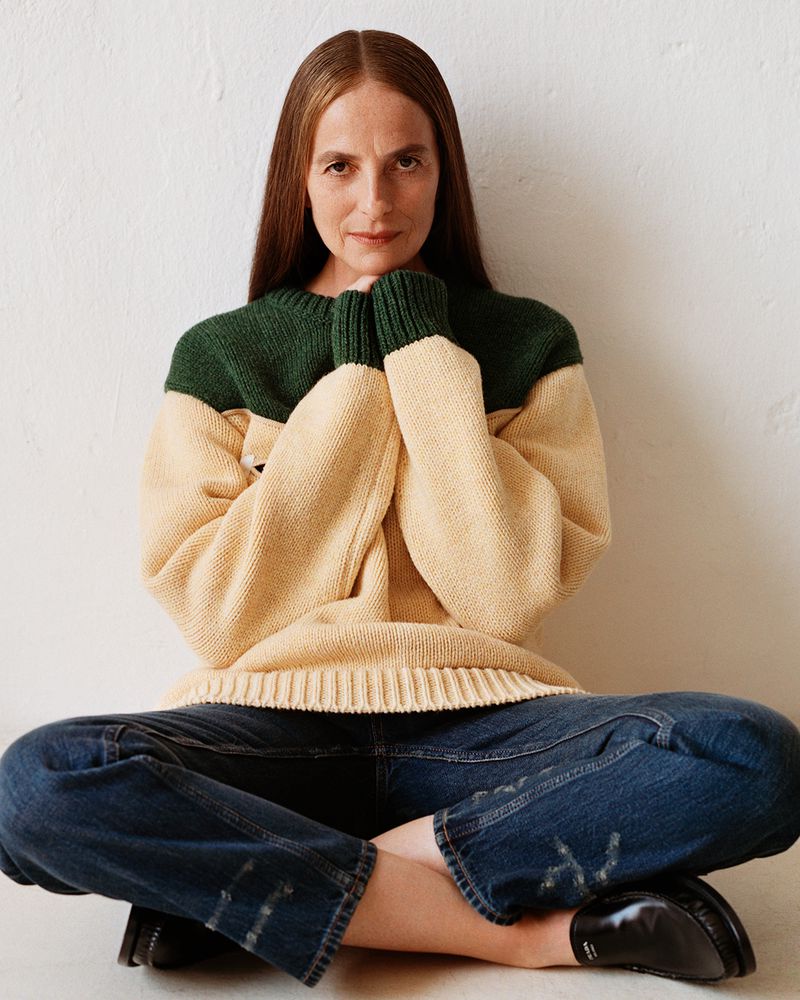 An adult woman with long brown hair is seated indoors, wearing a color-block sweater, jeans, and loafers, against a neutral wall.