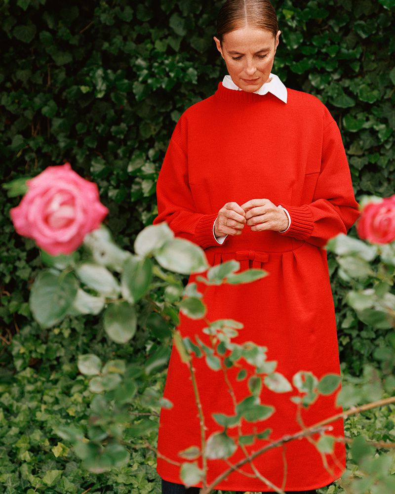 Adult woman in a red dress standing outdoors surrounded by roses and green foliage, looking down in natural daylight.
