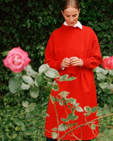 Adult woman in a red dress standing outdoors surrounded by roses and green foliage, looking down in natural daylight.