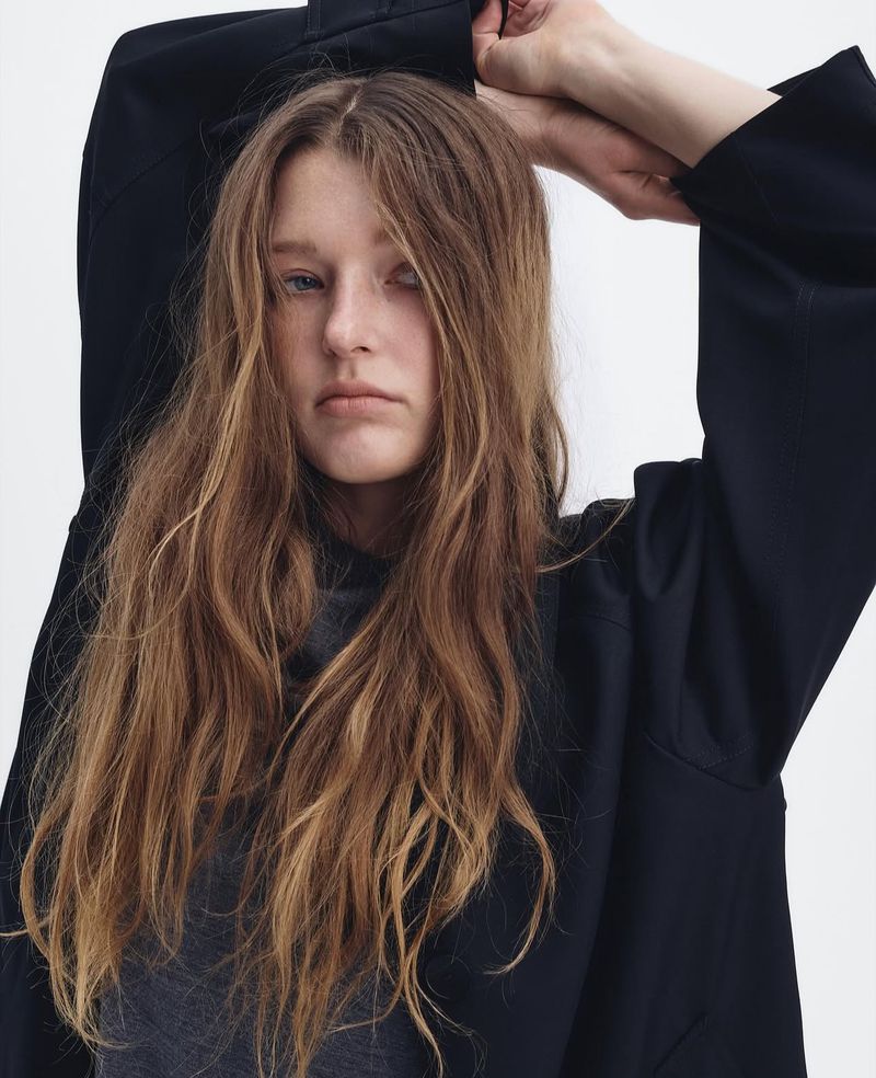Woman with long brown hair wearing a navy oversized jacket in a studio setting, striking a casual pose against a white backdrop.