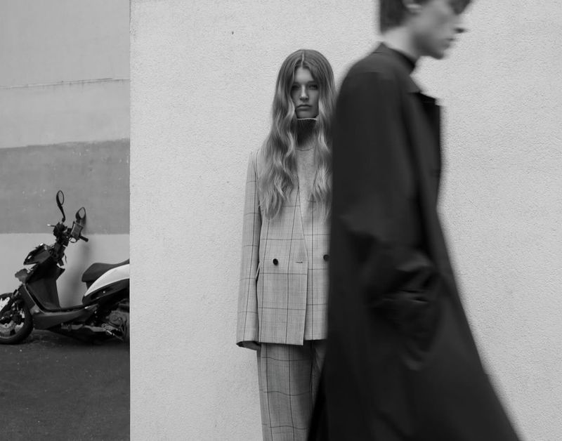 Two adults in monochrome photograph. Woman in plaid suit stands as man walks past in blurred motion. Parked scooter in background.