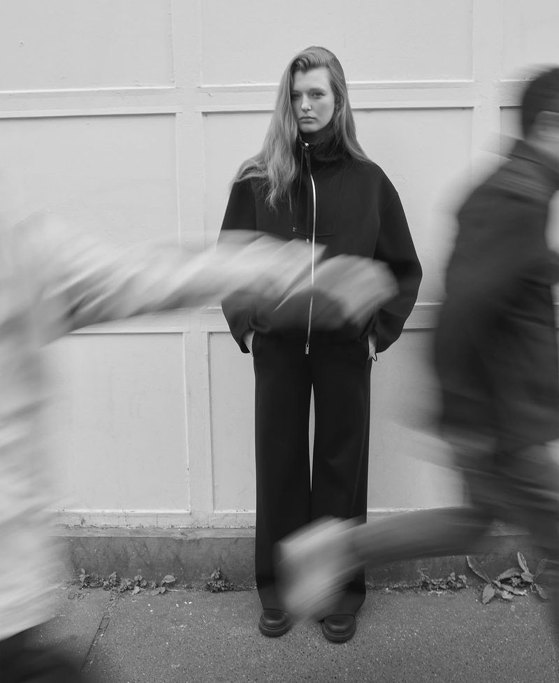 Monochrome fashion shot of a woman in a black jacket and pants posing against a light-colored wall with blurred figures in motion.