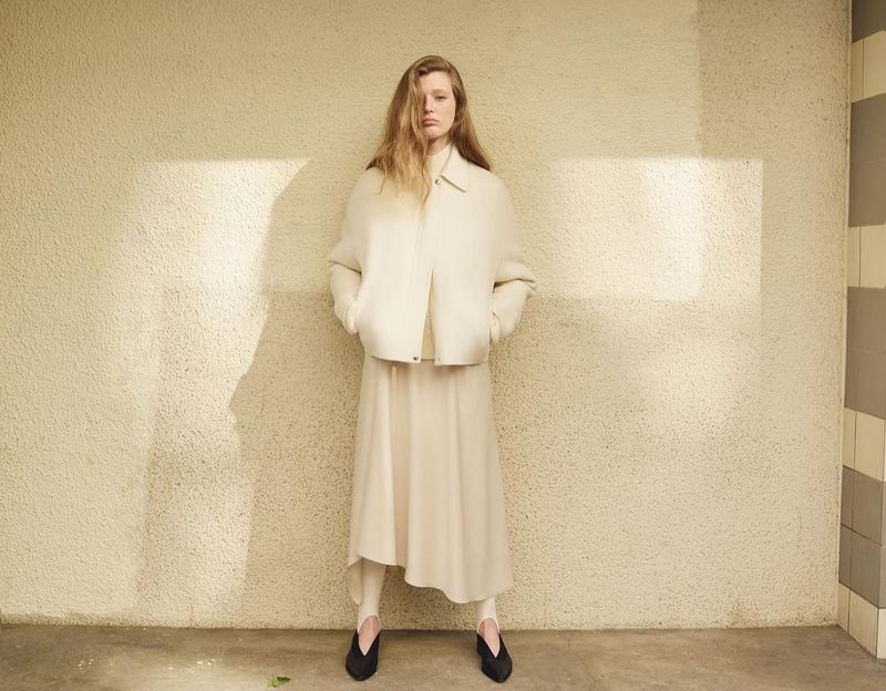 A woman in a cream-colored outfit stands indoors, posing against a textured wall with shadows in a fashion editorial-style photograph.