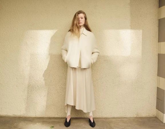 A woman in a cream-colored outfit stands indoors, posing against a textured wall with shadows in a fashion editorial-style photograph.