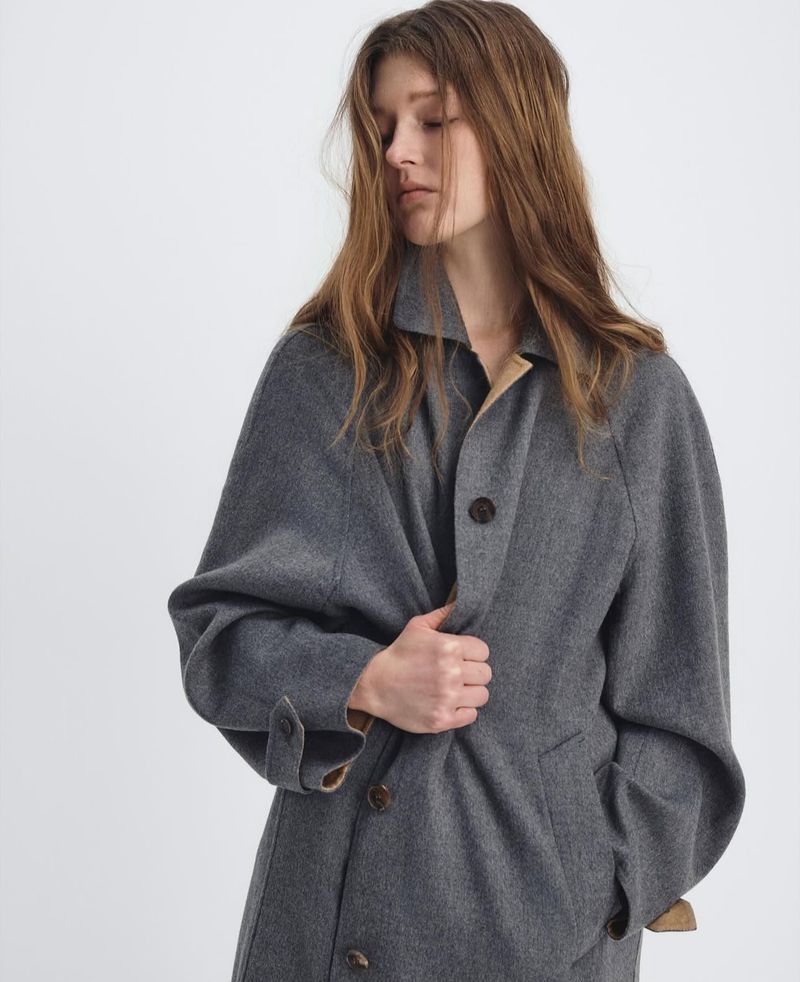 Adult female wearing a gray wool overcoat in a minimalist studio. Mid-shot, neutral facial expression, and soft studio lighting.
