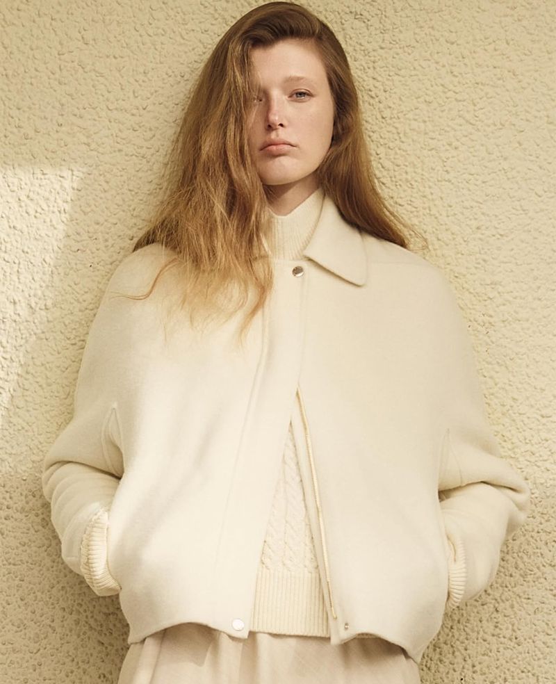 Blonde woman in a cream jacket and top stands against a textured wall, posing for a fashion portrait in a studio with soft lighting.