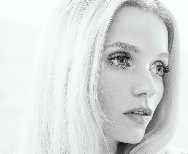 Monochromatic portrait of a fair-skinned adult woman with long blonde hair, looking off to the side against a blurred background.