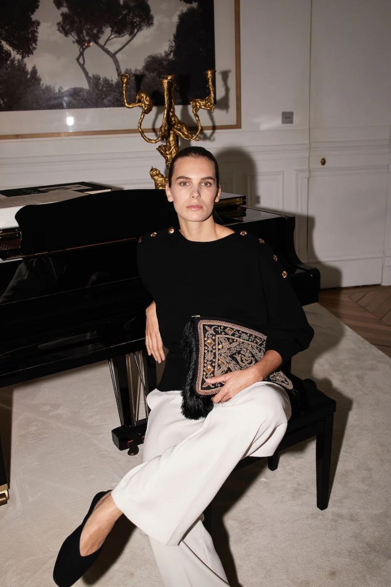 A fashionable adult female sits in an indoor setting with a piano and decorative candelabra, wearing a black sweater and white pants.