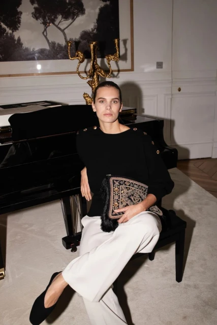 A fashionable adult female sits in an indoor setting with a piano and decorative candelabra, wearing a black sweater and white pants.
