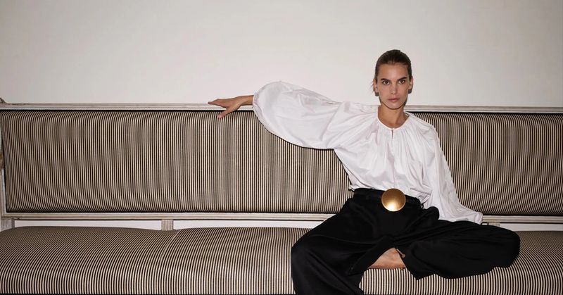 Adult woman in a white blouse and black pants sitting on a striped couch in a photo studio setting, looking at the camera.