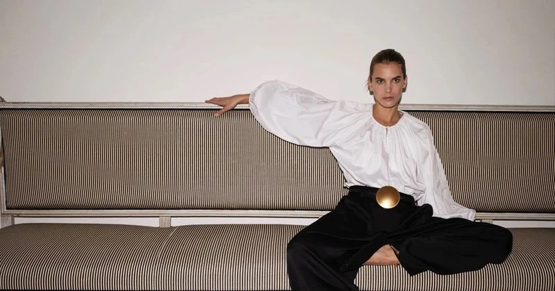 Adult woman in a white blouse and black pants sitting on a striped couch in a photo studio setting, looking at the camera.