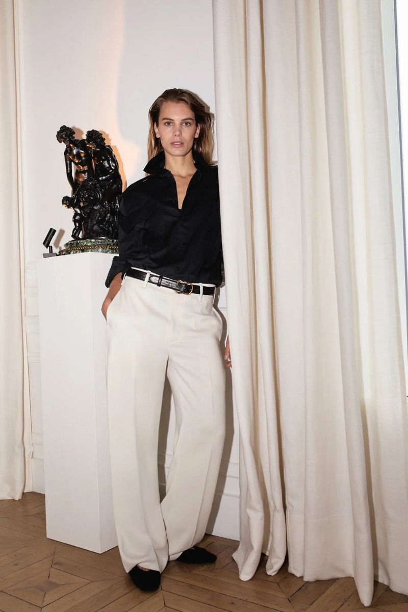 Elegant woman in a black blouse and white wide-leg pants stands next to a vintage sculpture and sheer curtains in a bright, indoor setting.