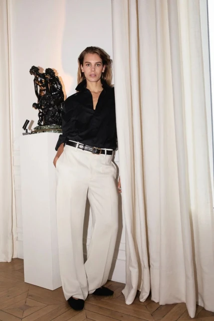 Elegant woman in a black blouse and white wide-leg pants stands next to a vintage sculpture and sheer curtains in a bright, indoor setting.