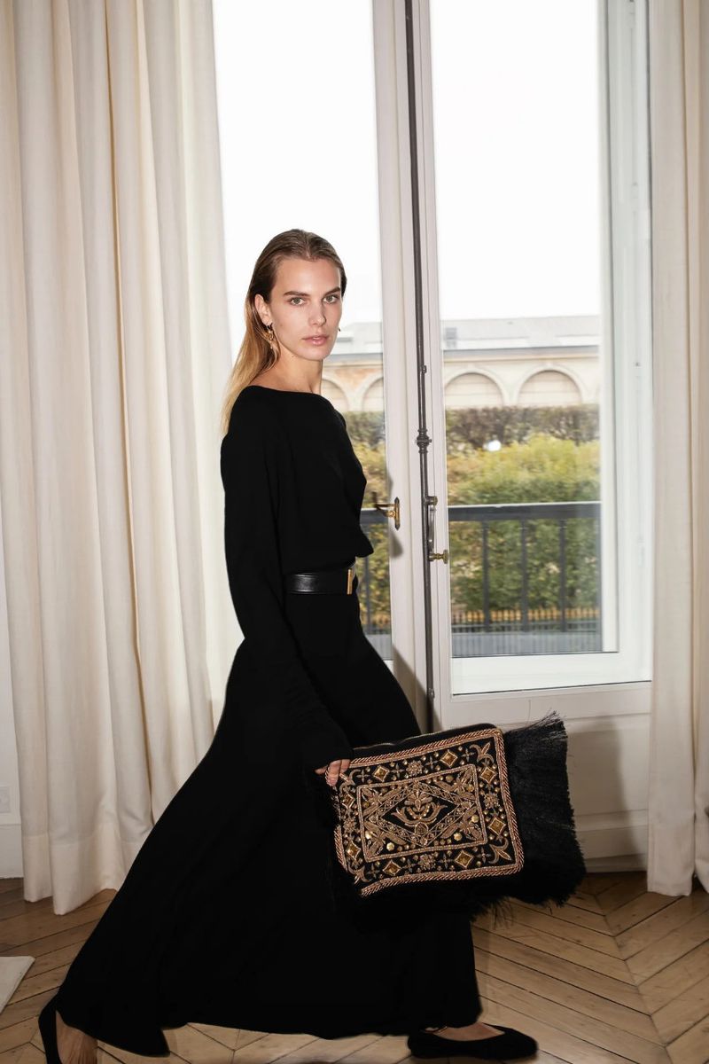 A female fashion model in a long black dress poses in front of a window with a beaded clutch. The scene is warmly lit and styled.