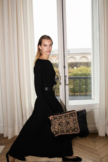 A female fashion model in a long black dress poses in front of a window with a beaded clutch. The scene is warmly lit and styled.
