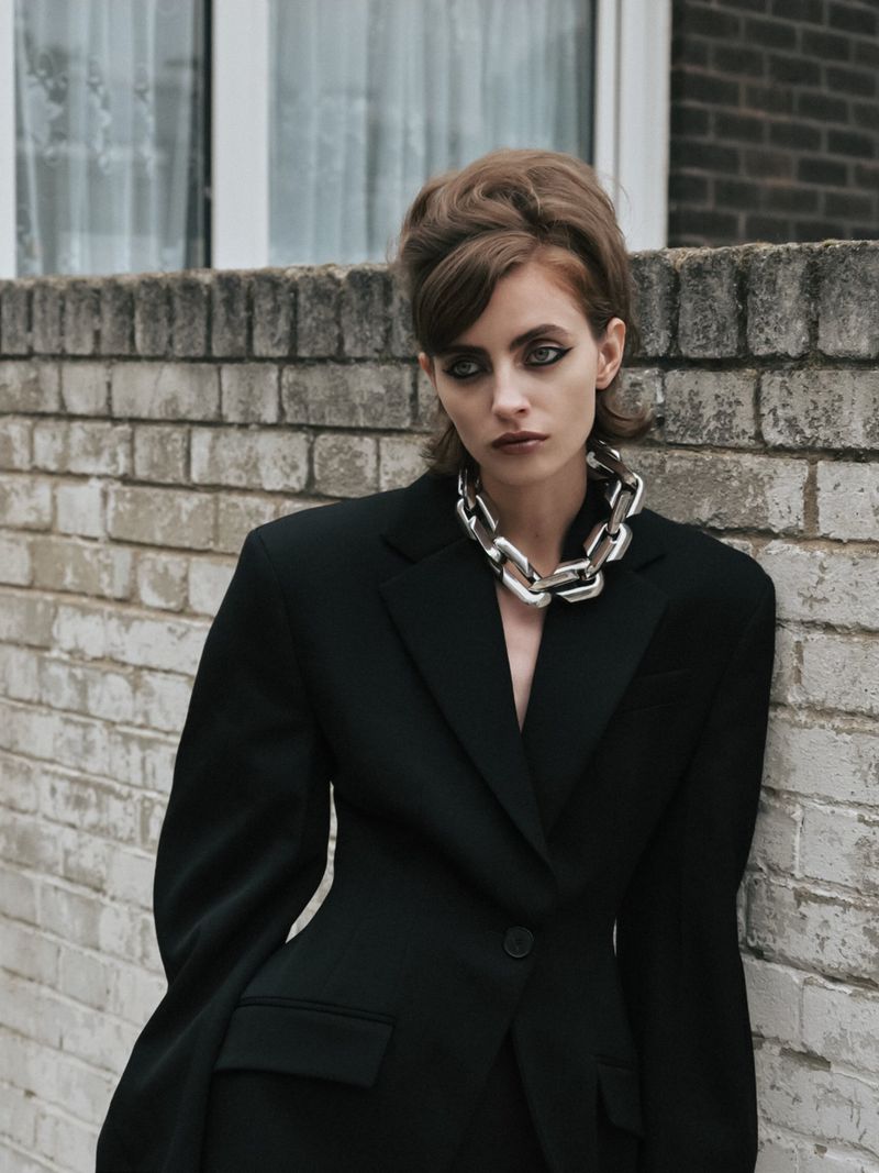 Portrait of a stylish adult woman in a black suit and bold chain necklace posing against a brick wall with a window in the background.