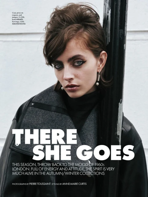 Close-up portrait of a young woman with a retro hairstyle wearing a grey coat and statement earrings, set against a neutral background.