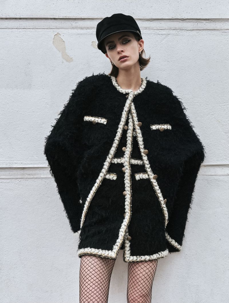 A stylish adult female model posing outdoors wearing a black tweed jacket, skirt, fishnet stockings, and cap against a wall backdrop.