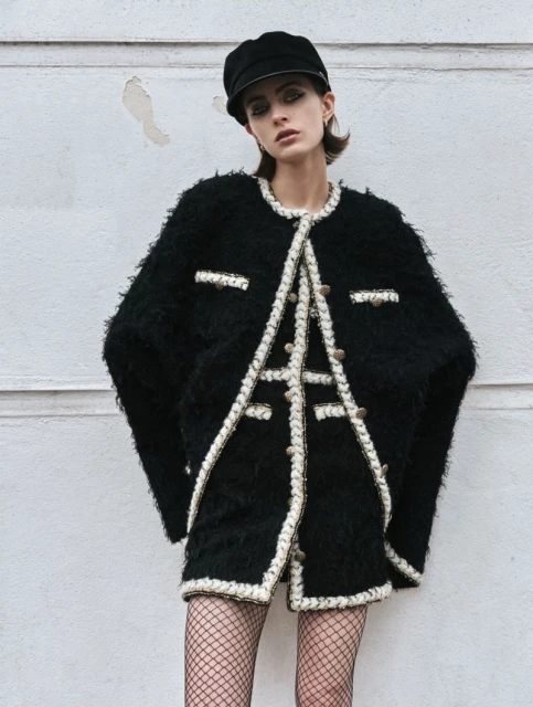 A stylish adult female model posing outdoors wearing a black tweed jacket, skirt, fishnet stockings, and cap against a wall backdrop.