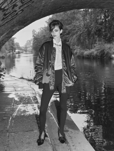 Black and white fashion shot of a female model in a jacket and leggings standing next to a canal, with a bridge in the background.