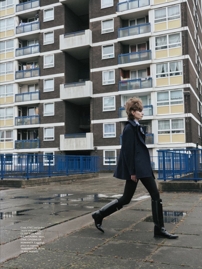 Fashion editorial image features a female fashion model in a dark coat and boots walking on a rooftop, with a tall building in the background.