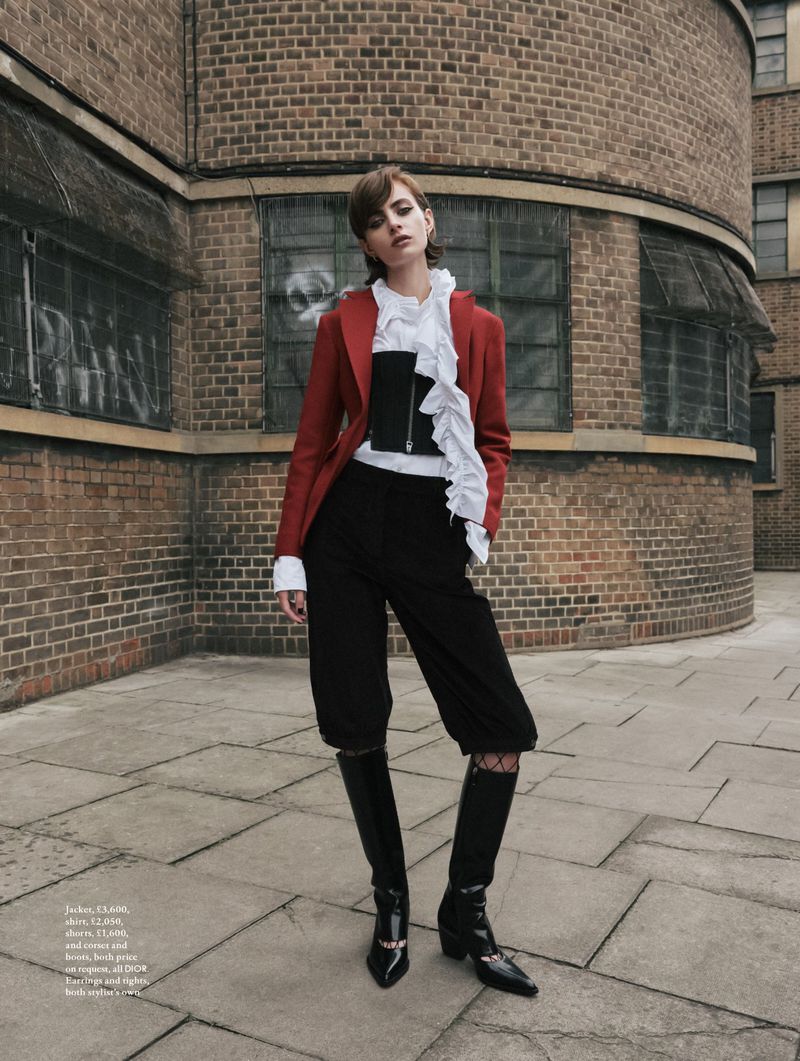 Fashionable woman in red blazer and black culottes poses confidently against brick architecture, showcasing edgy, modern style.
