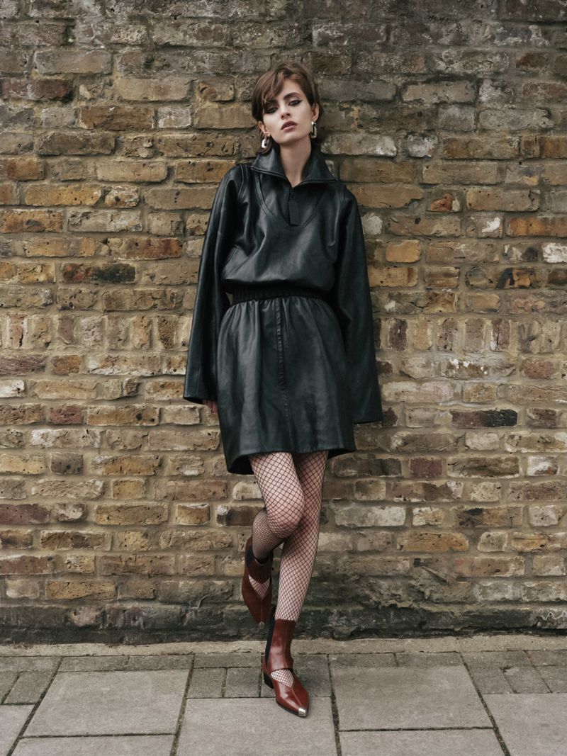 Adult woman in black mini dress poses against a brick wall outdoors. She wears fishnet tights and brown boots, in a street setting.