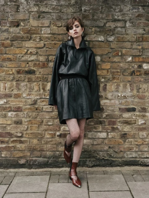 Adult woman in black mini dress poses against a brick wall outdoors. She wears fishnet tights and brown boots, in a street setting.
