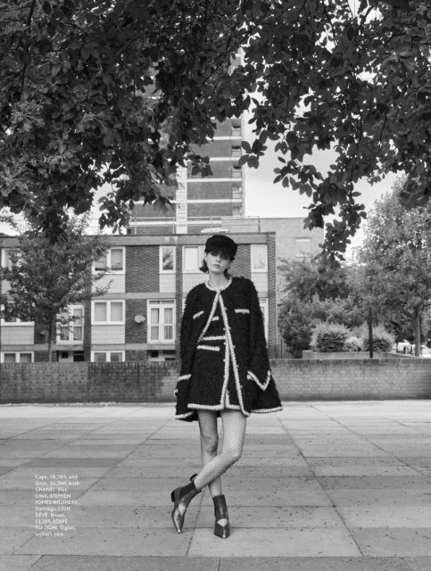 A fashion model poses outdoors wearing a black and white jacket, skirt, hat, fishnet stockings, and ankle boots against a cityscape backdrop.