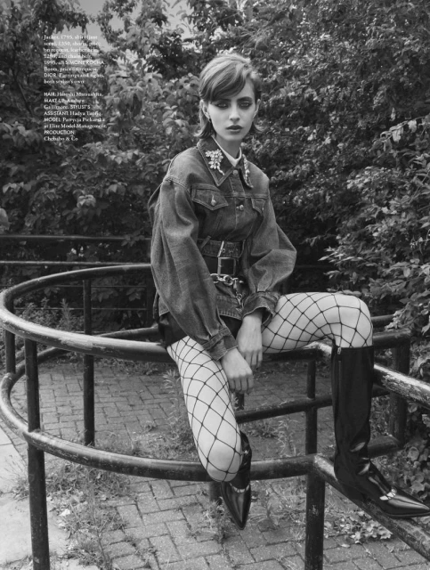Young woman poses in a denim jacket, fishnet tights, and boots on metal railing with trees in the background in a monochrome portrait.